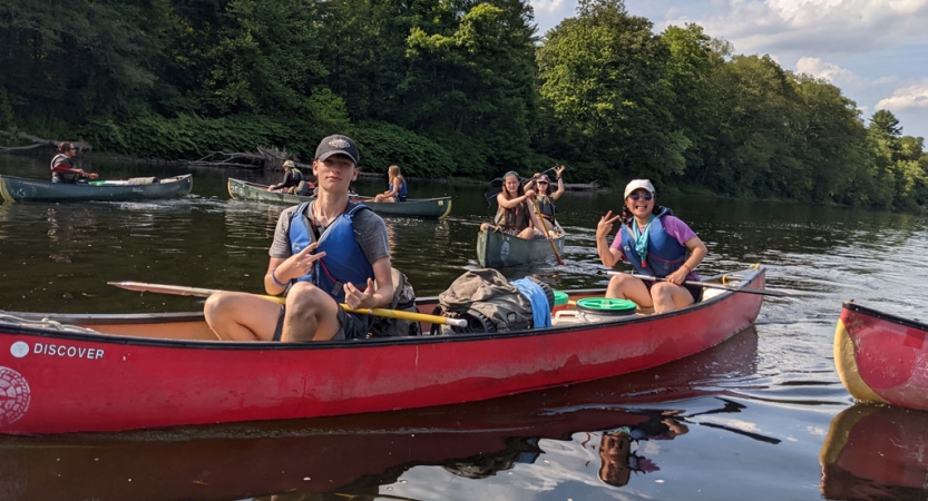 In the foreground, two people wearing life jackets sitting in a red canoe give the camera peace signs. There are people in other canoes in the background. 